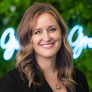 headshot of Angela Kujava, managing director of the Desai accelerator. Greenery backgroud with neon sign that reads "Go Blue » Grow Blue"